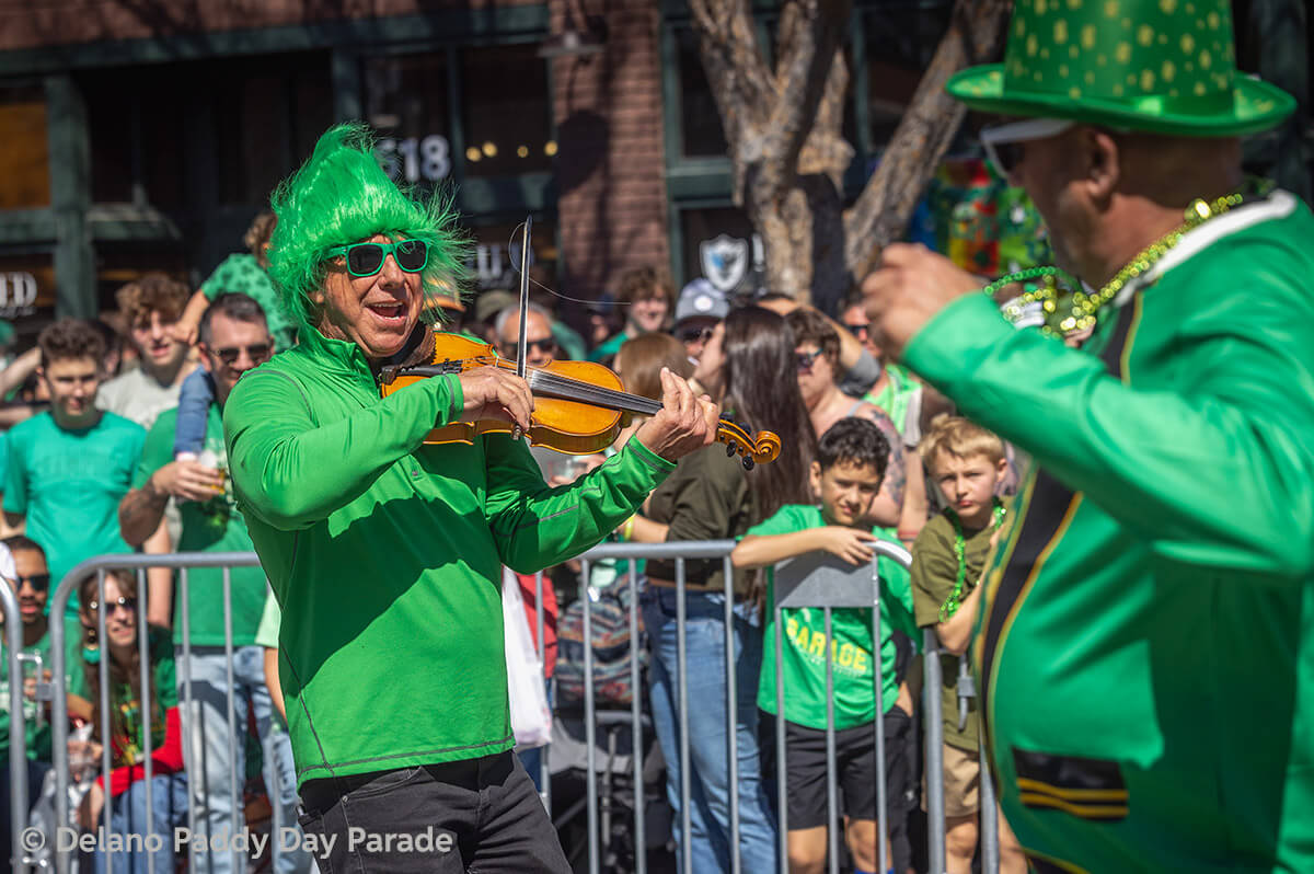 A man in a green wig plays the fiddle while another man dances.